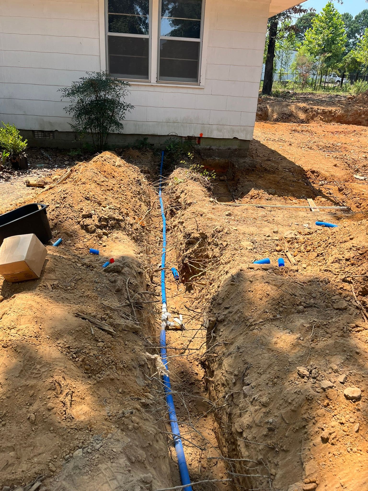 Trenches with blue pipes near a house foundation, prepared for plumbing installation.