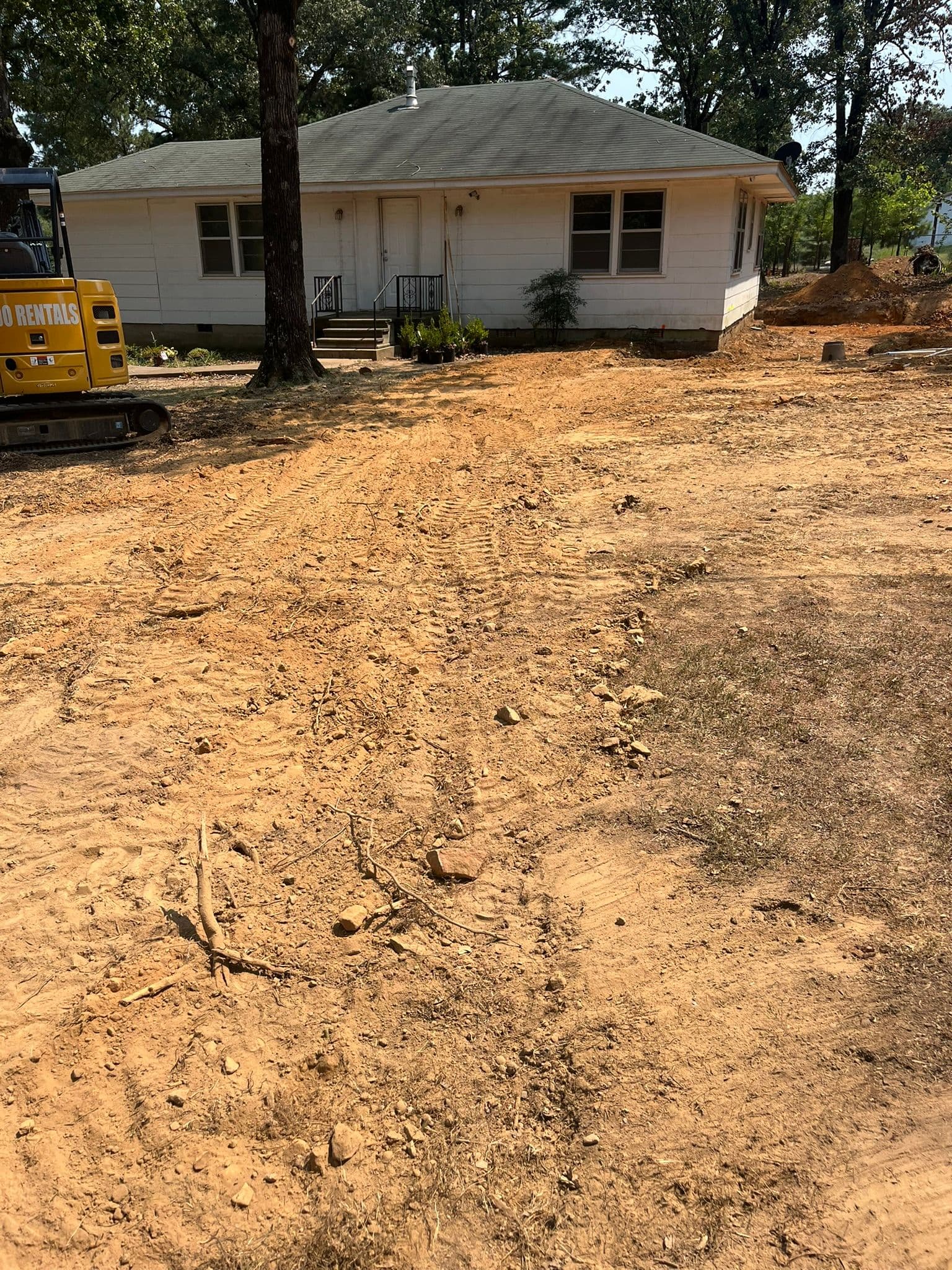 Construction site with a single-story white house, dirt, tire tracks, and machinery in view.