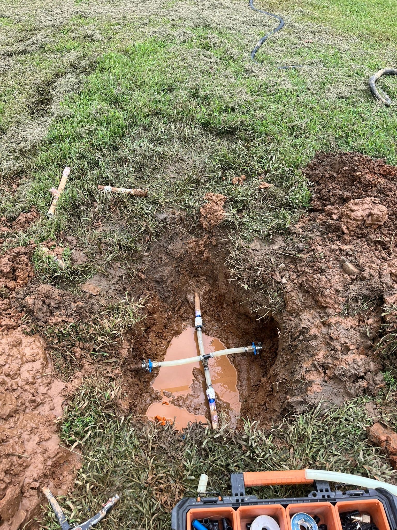 Exposed plumbing with water accumulation in a dug-out area of grass.